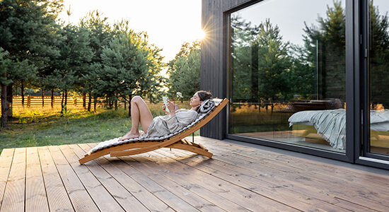 kobieta odpoczywająca na tarasie na drewnianym leżaku z książką w ręku na , w oddali zachód słońca i zagajnik 