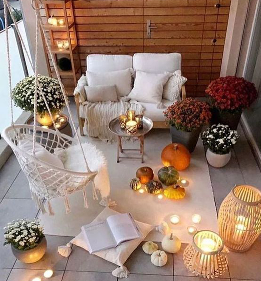 wygodna sofa na balkonie w bloku, przykryta ciepłym wełnianym kocem i obłożona puchatymi białymi poduszkami, krzesło brazylijskie z puchatymi poduszkami, na podłodze doniczki z roślinami, różnej wielkości dynie, odpalone w drewnianych lampionach świeczki