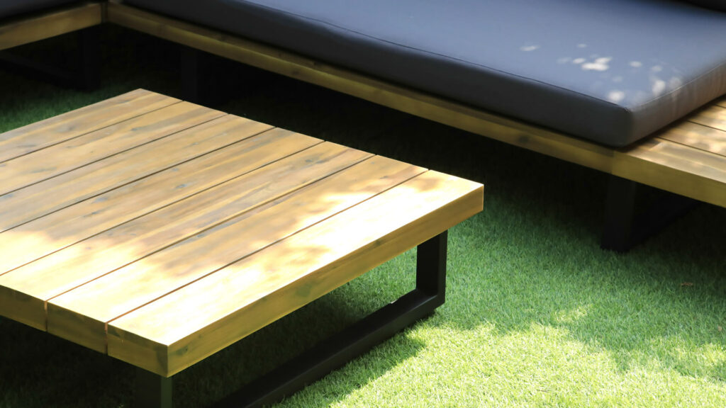 drewniany stolik ogrodowy z narożnikiem z szarymi poduszkami ustawiony na zielonej trawie w ogrodzie 