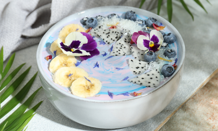  Pyszna miska na smoothie ze świeżymi owocami, jagodami i kwiatami na stole z kolorową teksturą. 