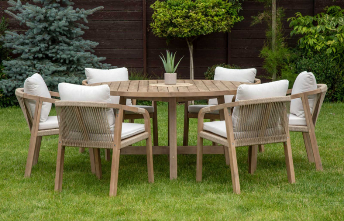 Okrągły stół z krzesłami Furrore Marigold 6+1 Round, drewniany z beżowymi poduszkami na siedziskach i oparaciach, zedstaw ustawiony na zielone trawie w ogrodzie 