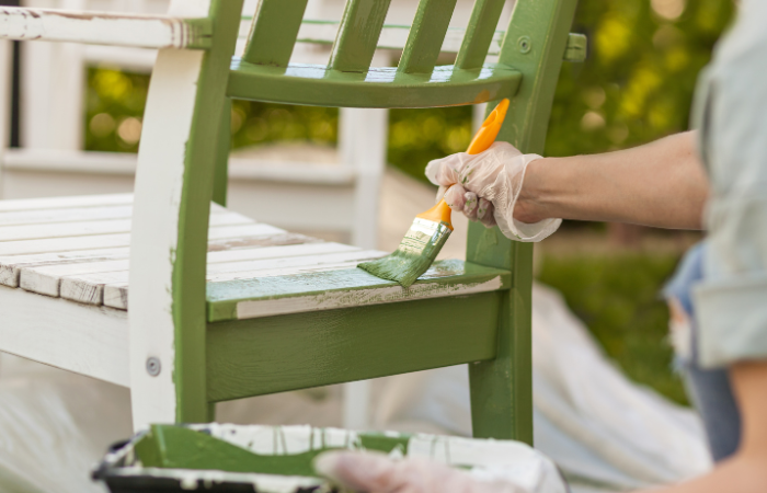 Malowanie krzesła pędzlem w rękawiczkach ochronnych. Pracownik maluje meble ogrodowe na zielono. Odnawianie, renowacja drewnianych mebli ogrodowych