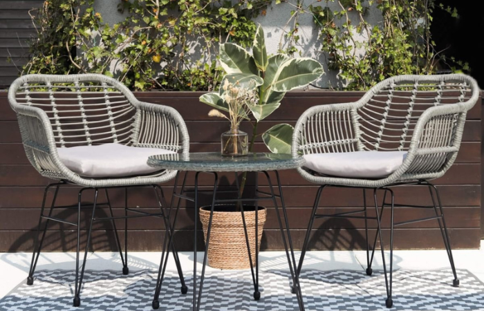 dwa metalowe krzesla z malym stolikiem ze szklanym blatem ustawione na balkonie wsrod skrzyń z roślinami, na podlodze szaro bialy dywan zewnetrzny 